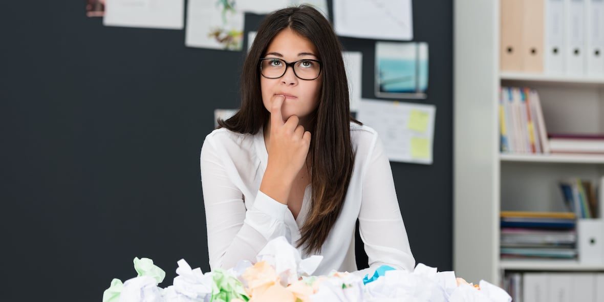 Young woman with writers block sitting in an office with a desk littered with crumpled paper as she sits looking thoughtfully into the air with her finger to her chin seeking new ideas